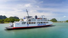 Ein fotorealistisches Bild einer großen blau-weißen Autofähre, die bei strahlendem Sonnenschein auf dem türkisblauen Meer in Richtung der grünen Hügel von Koh Samui zusteuert. Foto: Seatran Ferry