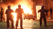 Irische Nationalpolizisten stehen in der Nähe eines brennenden Polizeiautos. Foto: Brian Lawless/Pa Wire/dpa