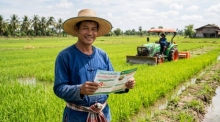Ein thailändischer Landwirt steht lächelnd auf seinem Reisfeld und hält eine Broschüre der BAAC-Bank in der Hand, im Hintergrund ein Traktor. Foto: Der Farang (ai)