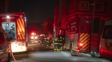 Feuerwehrleute stehen für Löscharbeiten auf einer Straße, nahe eines brennenden Gebäudes. Im Zentrum der Stadt sind bei einem Feuer in einem Gebäude mindestens 58 Menschen ums Leben gekommen. Foto: Uncredited/Ap/dpa