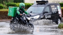 Ein Zusteller auf einem Motorrad fährt bei Regen an einem Auto vorbei. Foto: EPA-EFE/Ali Haider