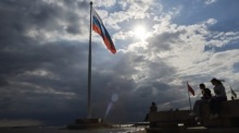Menschen spazieren an einer Uferpromenade in St. Petersburg entlang. Im Hintergrund sind große Flaggen Russlands und der ehemaligen Sowjetunion zu sehen. Foto: epa/Anatoly Maltsev