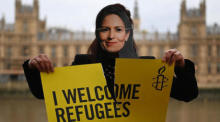 Ein Aktivist von Amnesty International, der eine Maske der britischen Innenministerin Priti Patel trägt, hält ein Plakat während einer "Refugees Welcome"-Demonstration in London. Foto: epa/Andy Rain