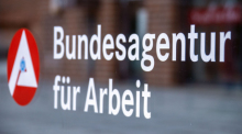 Das Hinweisschild für die Bundesagentur für Arbeit ist an einer Glasfläche vor einem Bürogebäude zu sehen. Foto: Jens Büttner/dpa-zentralbild/dpa
