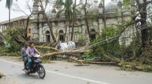 Einheimische fahren mit dem Motorrad auf einer Straße, während umgestürzte Bäume in der Nähe einer Moschee nach dem Zyklon «Mocha» liegen. Foto: Uncredited/dpa
