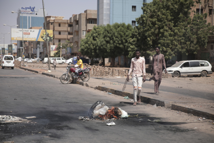 Personen gehen auf dem Fußgängerweg einer Straße entlang, auf der Brandspuren zu sehen sind und ein Müllsack brennt. Foto: Marwan Ali/Ap/dpa