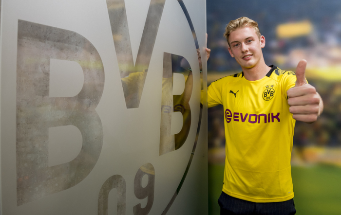 Neuzugang Julian Brandt steht neben einem großen Logo vom BVB. Foto: Alexandre Simoes/Bvb/Borussia Dortmund GmbH & Co/dpa 