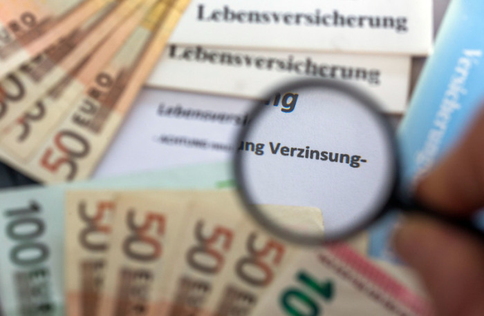 Versicherungsunterlagen liegen zwischen Euro-Geldscheinen und durch eine Lupe ist der Begriff «Verzinsung» zu sehen. Foto: Jens Büttner/Dpa-zentralbild/dpa