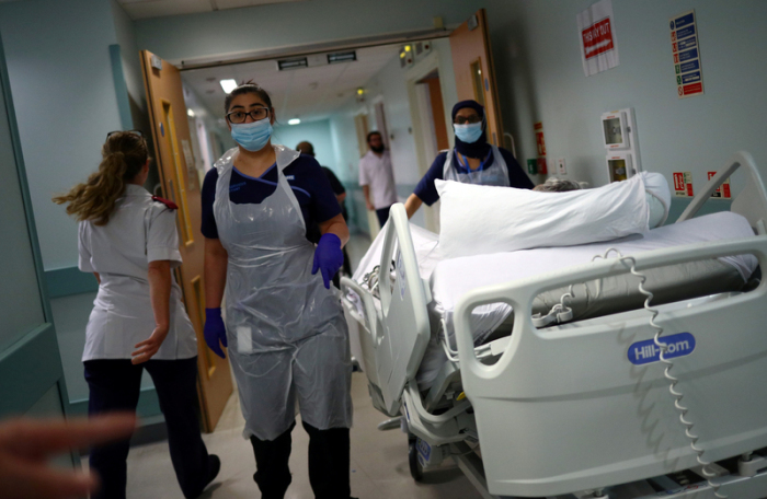 Medizinische Mitarbeiter des Royal Blackburn Lehrkrankenhaus in Schutzkleidung schieben einen Patienten auf einem Krankenhausbett durch einen Korridor. Foto: Hannah Mckay/Pa Wire/dpa