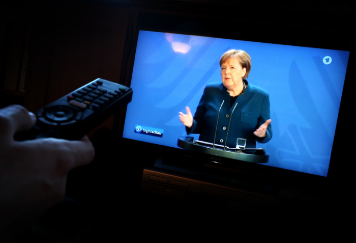 Ein junger Mann verfolgt in einem Wohnzimmer die Übertragung einer Ansprache der Bundeskanzlerin Angela Merkel (CDU) zur Lage im Zusammenhang mit dem Coronavirus. Foto: Karl-Josef Hildenbrand/Dpa