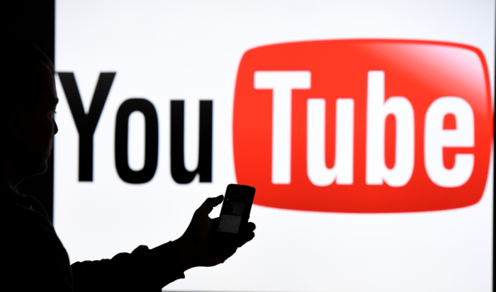 Ein Mann mit Smartphone steht vor dem Logo der Internet-Videoplattform Youtube. Seit anderthalb Jahrzehnten gibt es Youtube. Foto: Carsten Rehder/Dpa