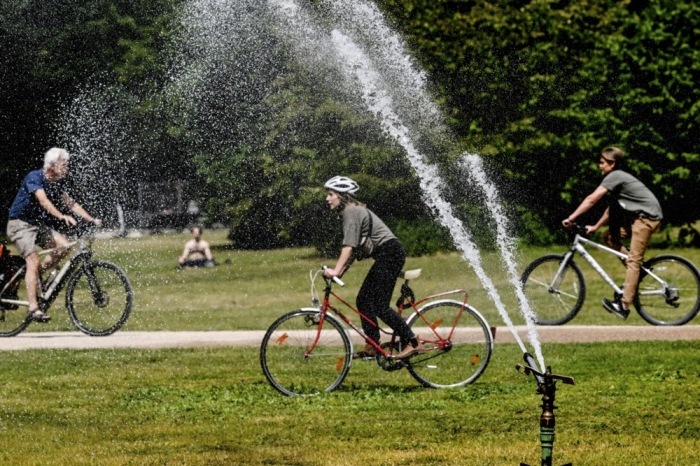 An einem warmen Tag im Tiergarten in Berlin fahren Radfahrer an einem Rasensprenger vorbei, der Wasser auf den Platz spritzt. Foto: epa/Filip Singer