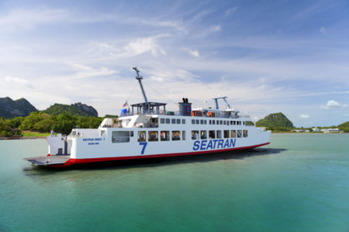 Ein fotorealistisches Bild einer großen blau-weißen Autofähre, die bei strahlendem Sonnenschein auf dem türkisblauen Meer in Richtung der grünen Hügel von Koh Samui zusteuert. Foto: Seatran Ferry