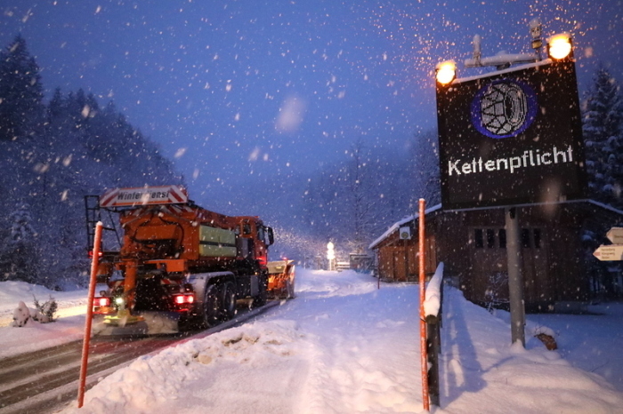 Ein Schild weist auf die Schneekettenpflicht am Riedbergpass im bayerischen Landkreis Oberallgäu hin, während daneben ein Räumfahrzeug vom Winterdienst den Schnee von der Straße räumt. Foto: Davor Knappmeyer/dpa