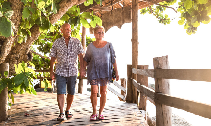 Ein älteres Paar genießt einen entspannten Spaziergang auf einem Holzsteg unter tropischen Bäumen. Foto: Adobe Stock/Mirko Vitali