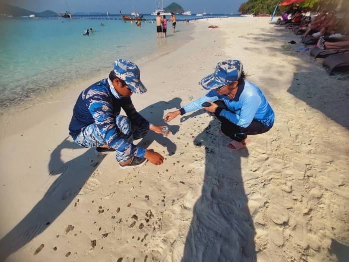 Einsatzkräfte sammeln Teerreste am Strand von Koh He ein, während Badegäste im Hintergrund weiter im Wasser sind. Foto: Newshawk Phuket