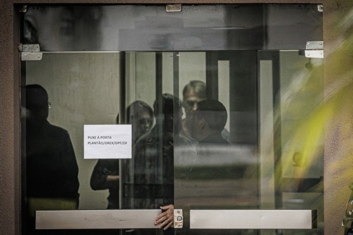 Jair Bolsonaro steht hinter einer Glastür im Polizeihauptquartier in Brasília, wo er nach Manipulation seiner elektronischen Fußfessel angehört wird. Foto: EPA/Isaac Fontana