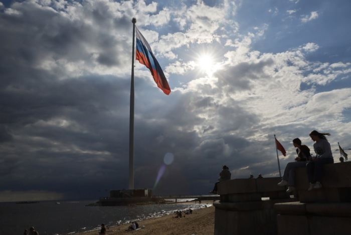 Menschen spazieren an einer Uferpromenade in St. Petersburg entlang. Im Hintergrund sind große Flaggen Russlands und der ehemaligen Sowjetunion zu sehen. Foto: epa/Anatoly Maltsev
