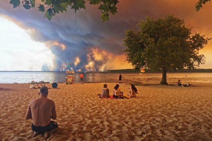 Strandbesucher sitzen am Strand von Moutchic in Lacanau im Südwesten Frankreichs, während Rauchwolken von einem Waldbrand in den Himmel aufsteigen. Foto: Maximilien Lamy/Afp/dpa