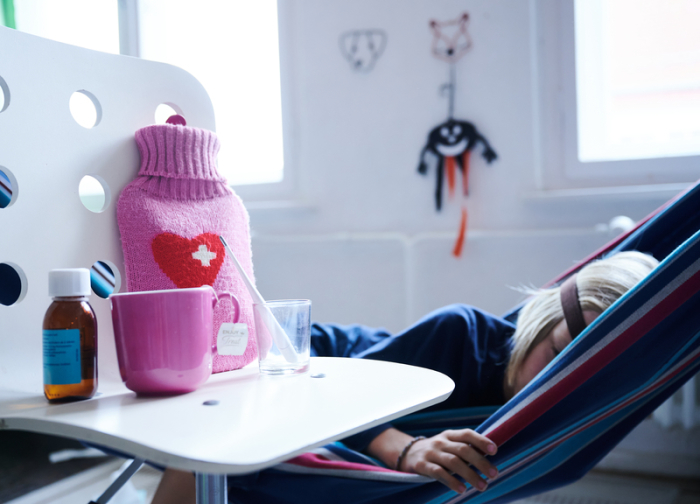 Fieberthermometer, Hustensaft, Wärmflasche und eine Tasse Tee stehen auf einem Stuhl, während ein Kind im Hintergrund in einer Hängematte liegt (gestellte Szene). Foto: Annette Riedl/dpa