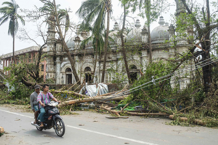 Einheimische fahren mit dem Motorrad auf einer Straße, während umgestürzte Bäume in der Nähe einer Moschee nach dem Zyklon «Mocha» liegen. Foto: Uncredited/dpa